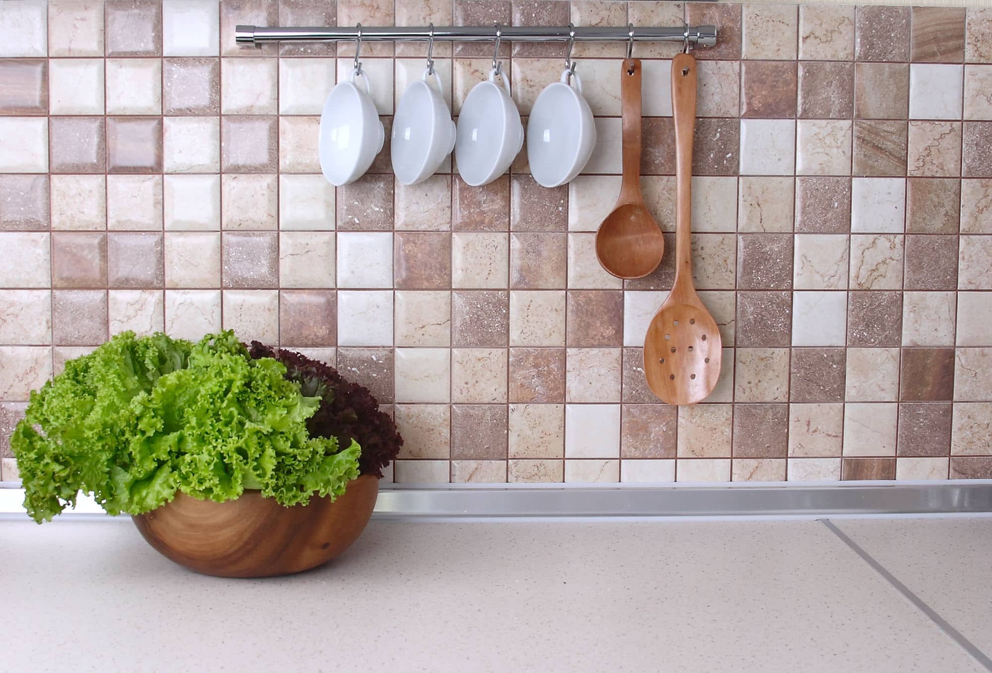 Arbeitsplatte mit Salatschüssel, dahinter Mosaikfliesen mit Hängeleiste für Tassen und Kochlöffel, dazwischen eine silberne Abschlussleiste