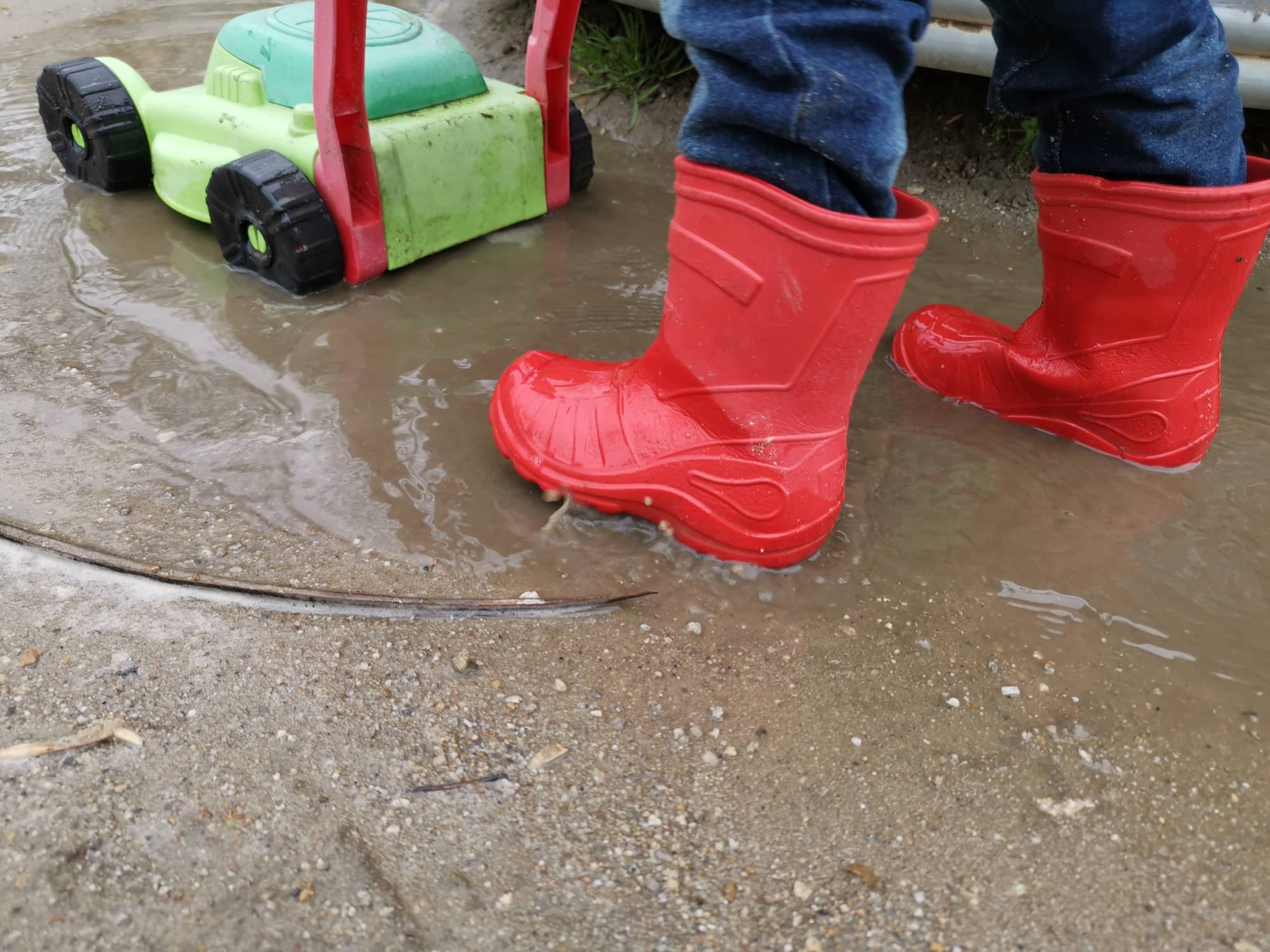 In der Nahaufnahme sieht man die Beine und Stiefel eines kleinen Kindes, das einen Spielzeugrasenmäher durch eine Matschpfütze schiebt.