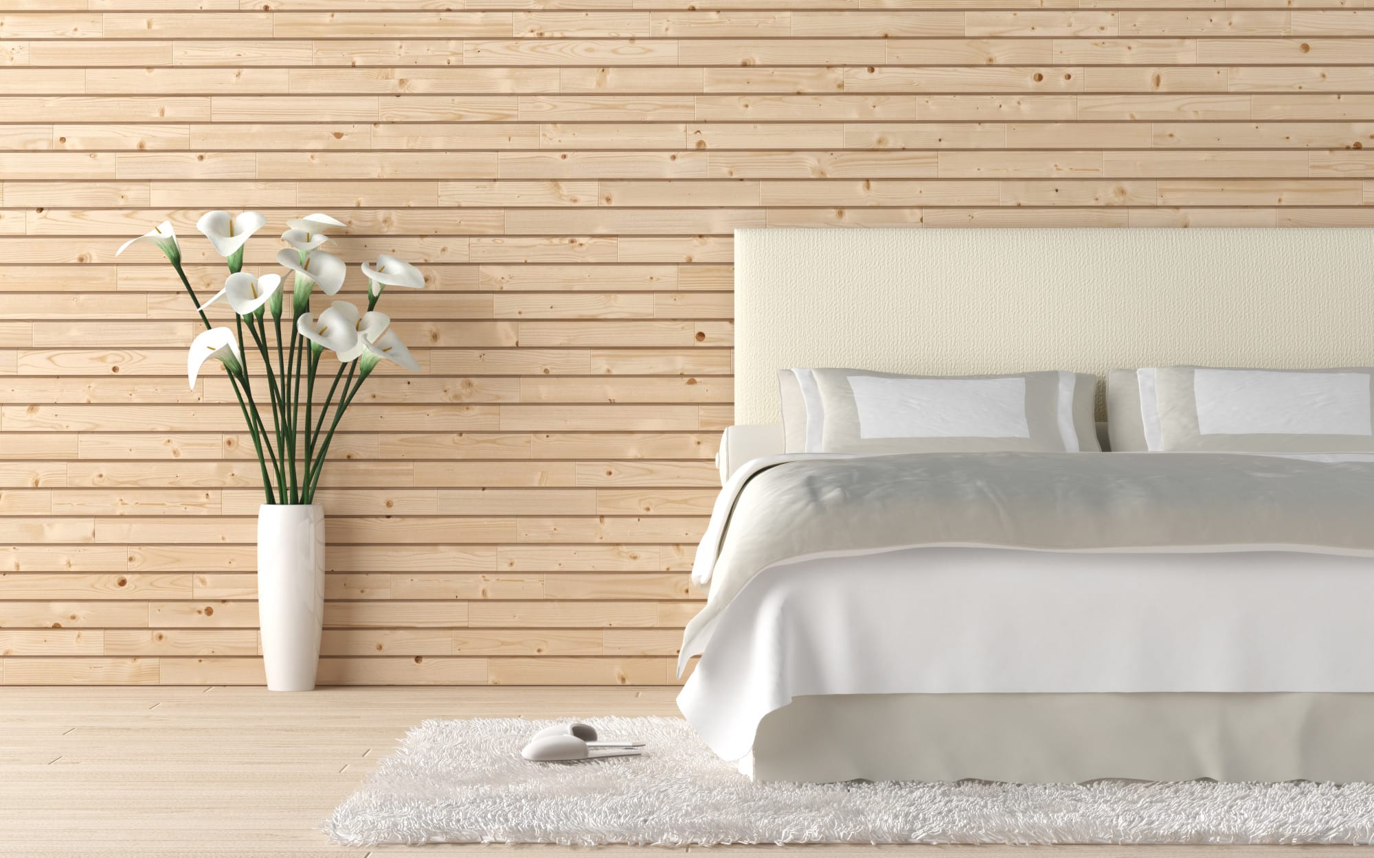 Ein weißes Bett mit grauer Tagesdecke steht im Schlafzimmer vor einer hellen Holzwand auf einem weißen Teppich und daneben steht eine Vase mit weißen Blumen.