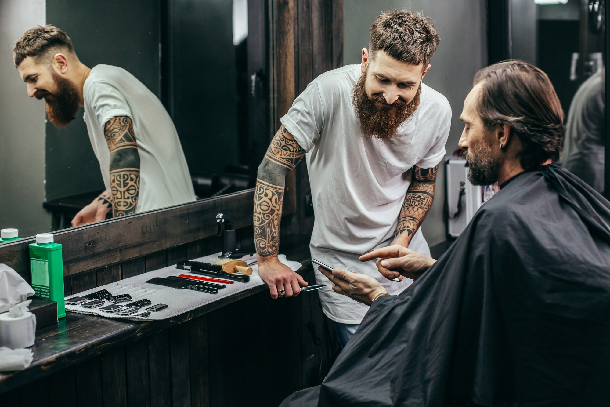 Friseur mit Bart und Tattoos schneidet einem Kunden mit schwarzem Umhang die Haare