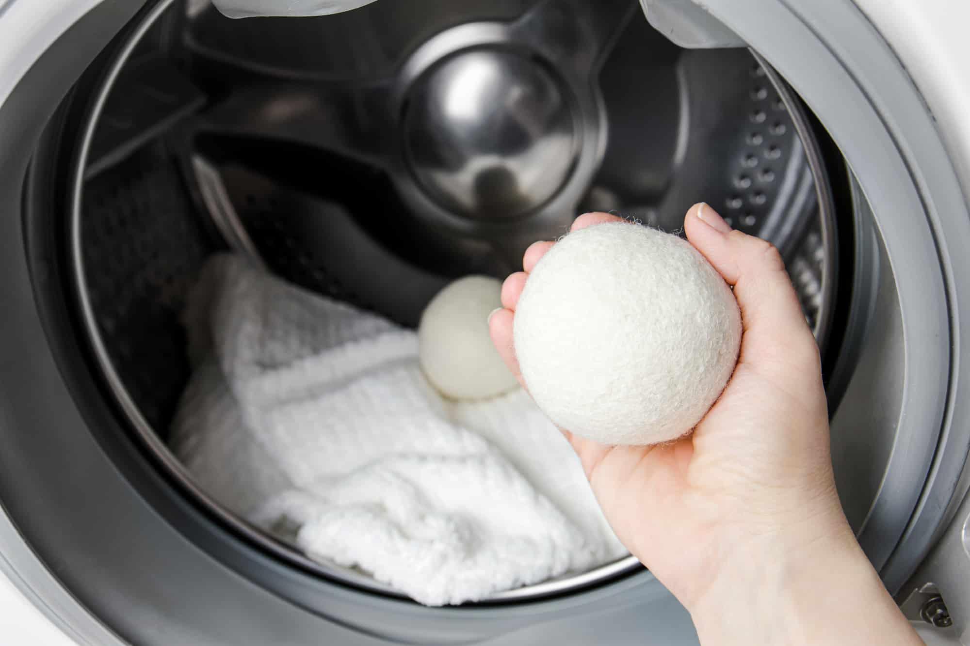 Hand hält einen weißen Wollball vor die geöffnete Trommel mit weißer Wäsche