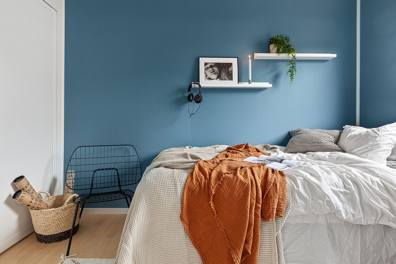 Ungemachtes Bett mit weißen Kissen und orangefarbener Decke vor blauer Wand