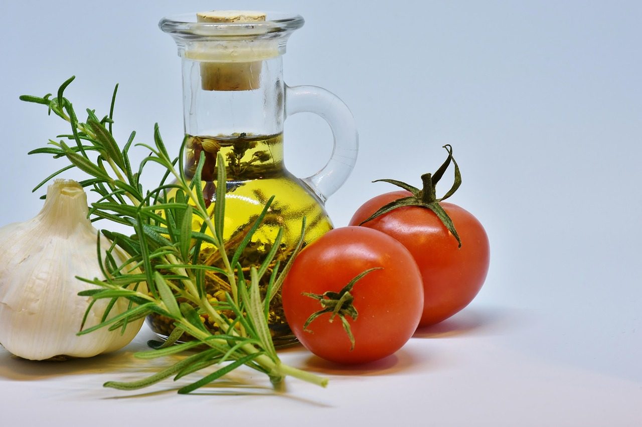 Glaskaraffe mit Speiseöl neben Tomaten, Knoblauch und Rosmarin